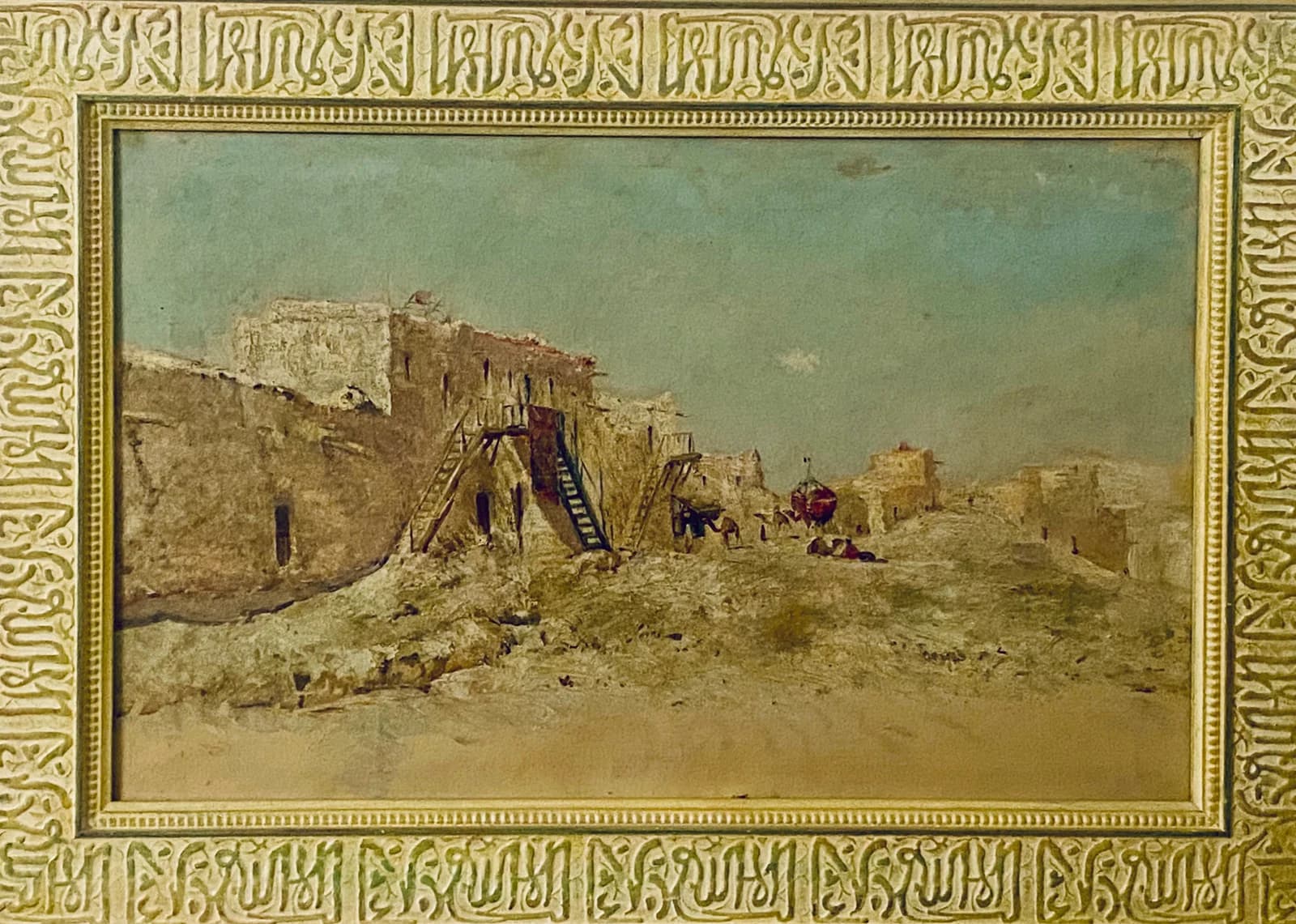 A reader's photograph of a landscape painting in a carved gilt frame: pale walls and towers of a fortified village under a hazy blue sky, a wooden ladder against the ramparts, sandy ground with small figures.